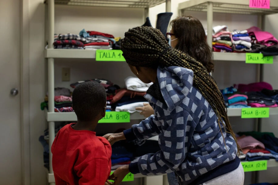 A volunteer helps a woman and boy pick out fresh clothes for the boy from two shelves of children's clothing.